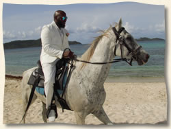 Groom in white suit on white horse riding to his beach wedding.